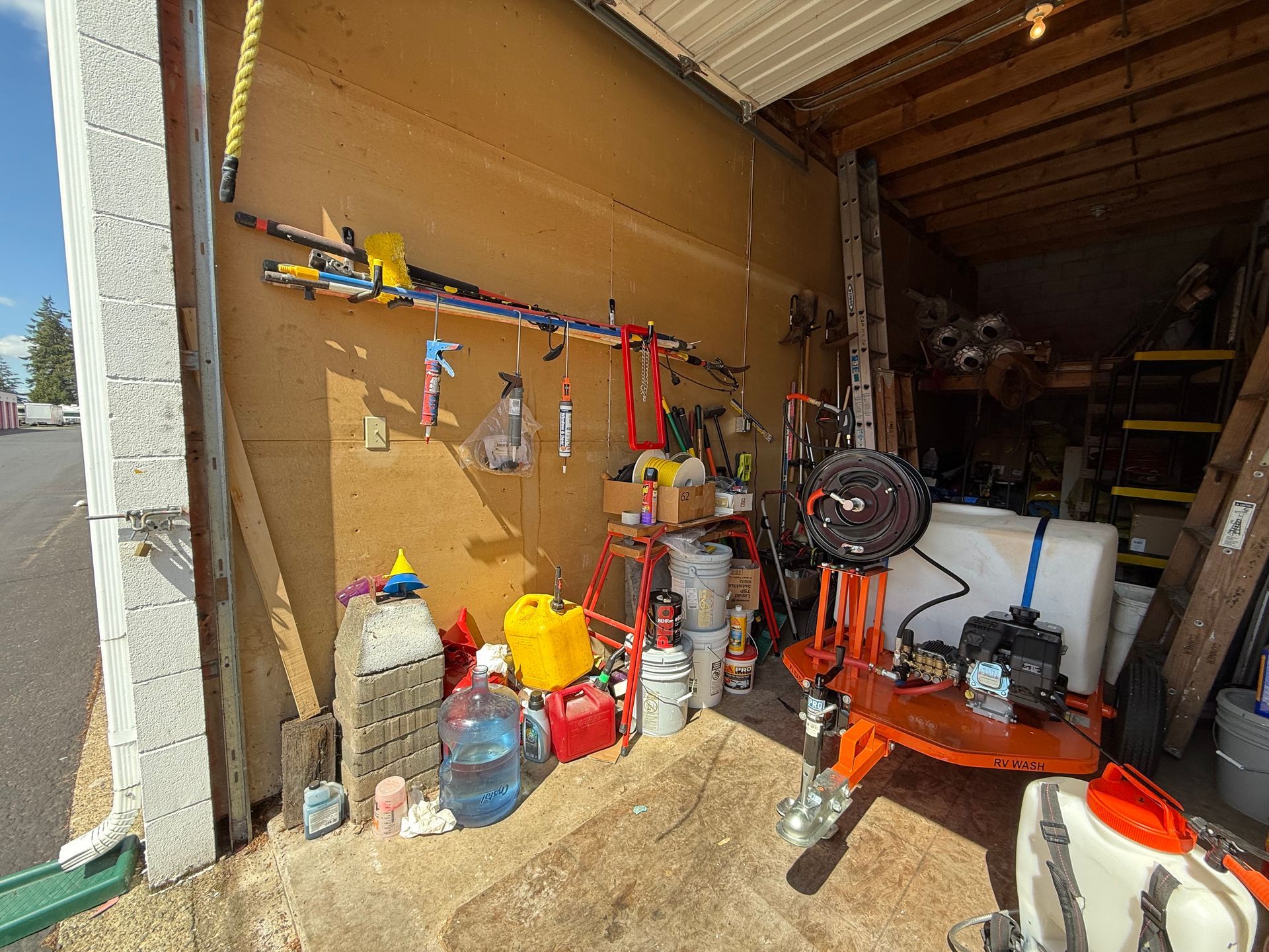 A cluttered storage area with tools, equipment, and supplies. Beige walls, concrete pillars, and a partly open door.