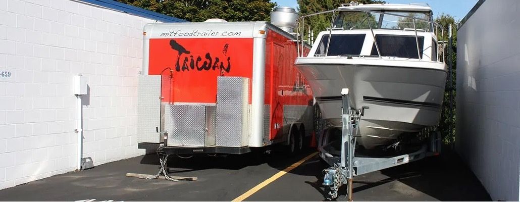 A boat on a trailer and a food trailer parked between two white walls. The food trailer is red and white.