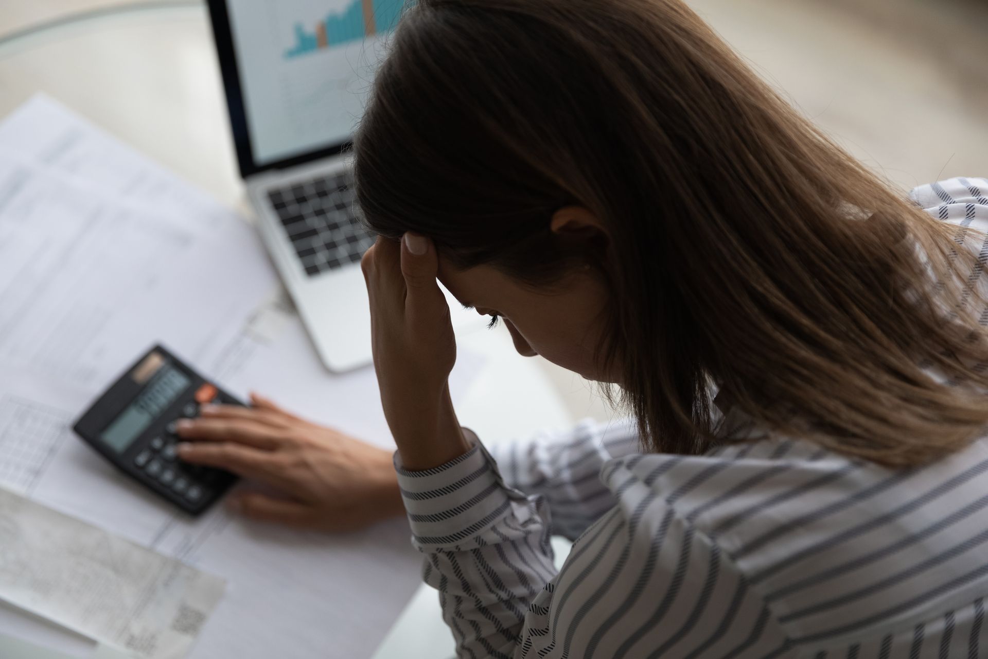 Person holding head while using calculator beside laptop and papers on a desk