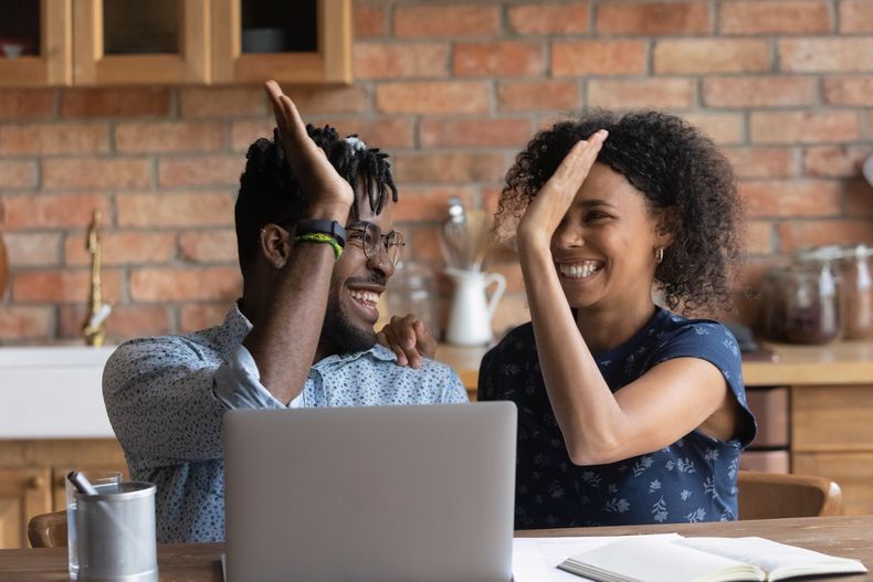 Two people high-five across a laptop in a café, smiling in a brick-walled setting.