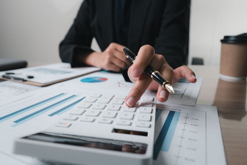 Person calculating finances with a pen, using a calculator on a desk with charts and documents