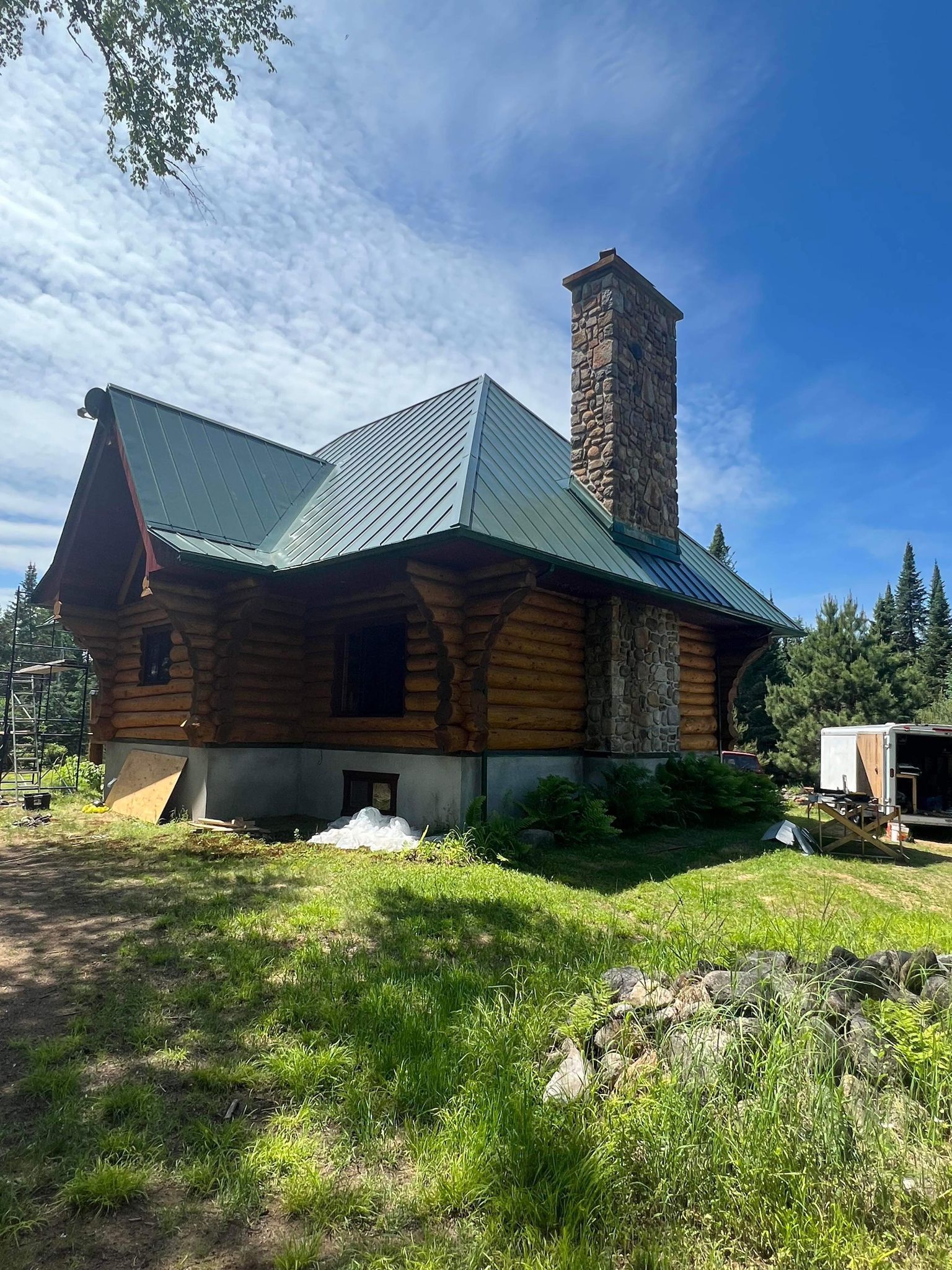 A log cabin with a green roof and a stone chimney is sitting in the middle of a grassy field.