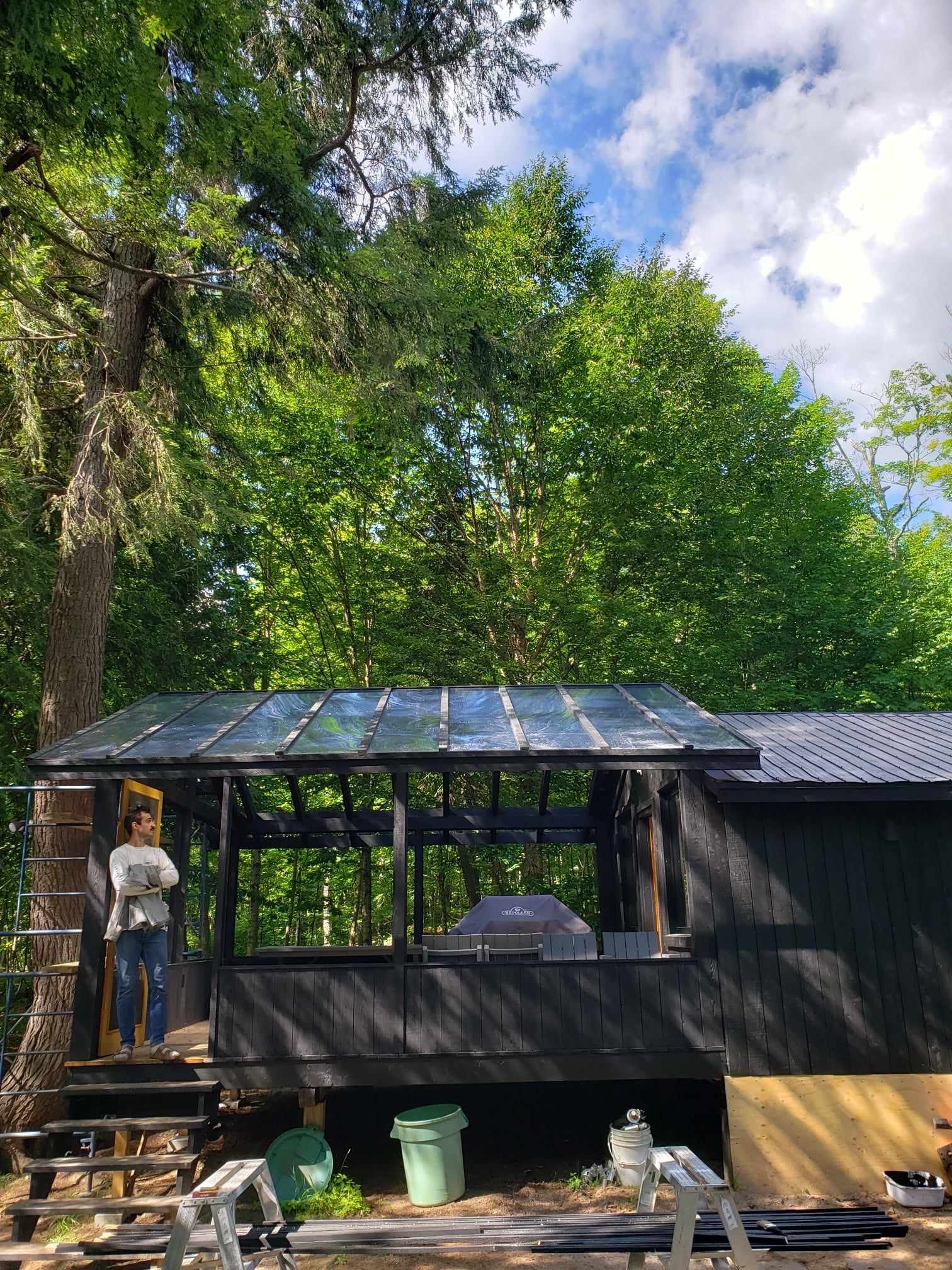 A woman is standing on a ladder in front of a black house surrounded by trees.