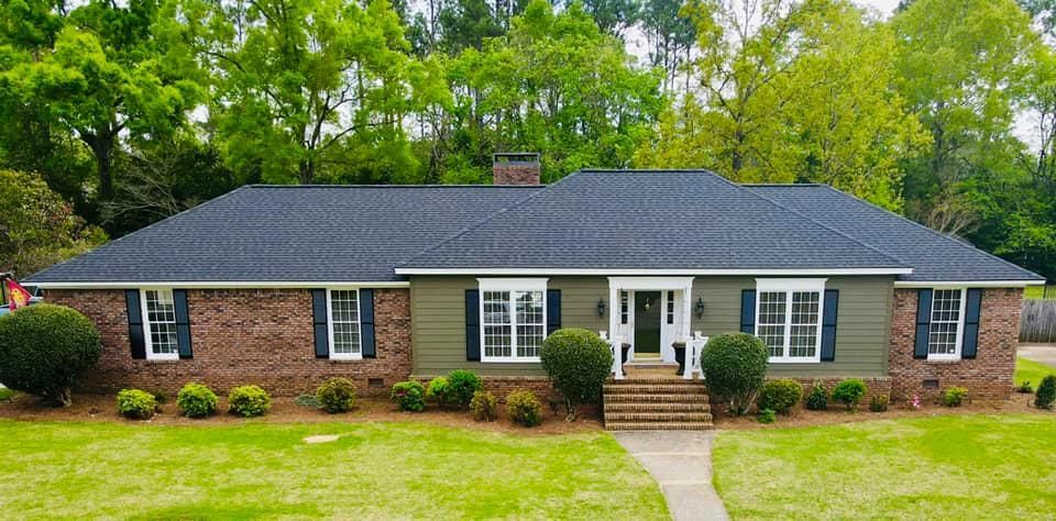 A single-story house with a black roof, brick and green siding, and a small front yard with bushes.