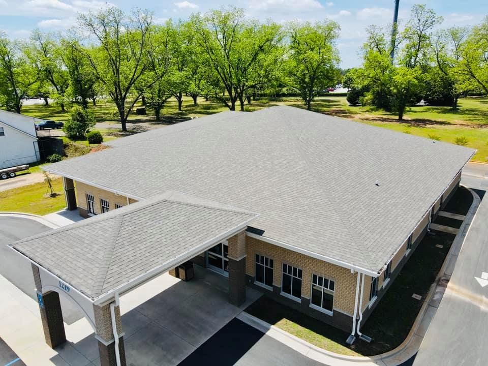 Exterior view of a one-story building with a light gray roof and awning, surrounded by trees.