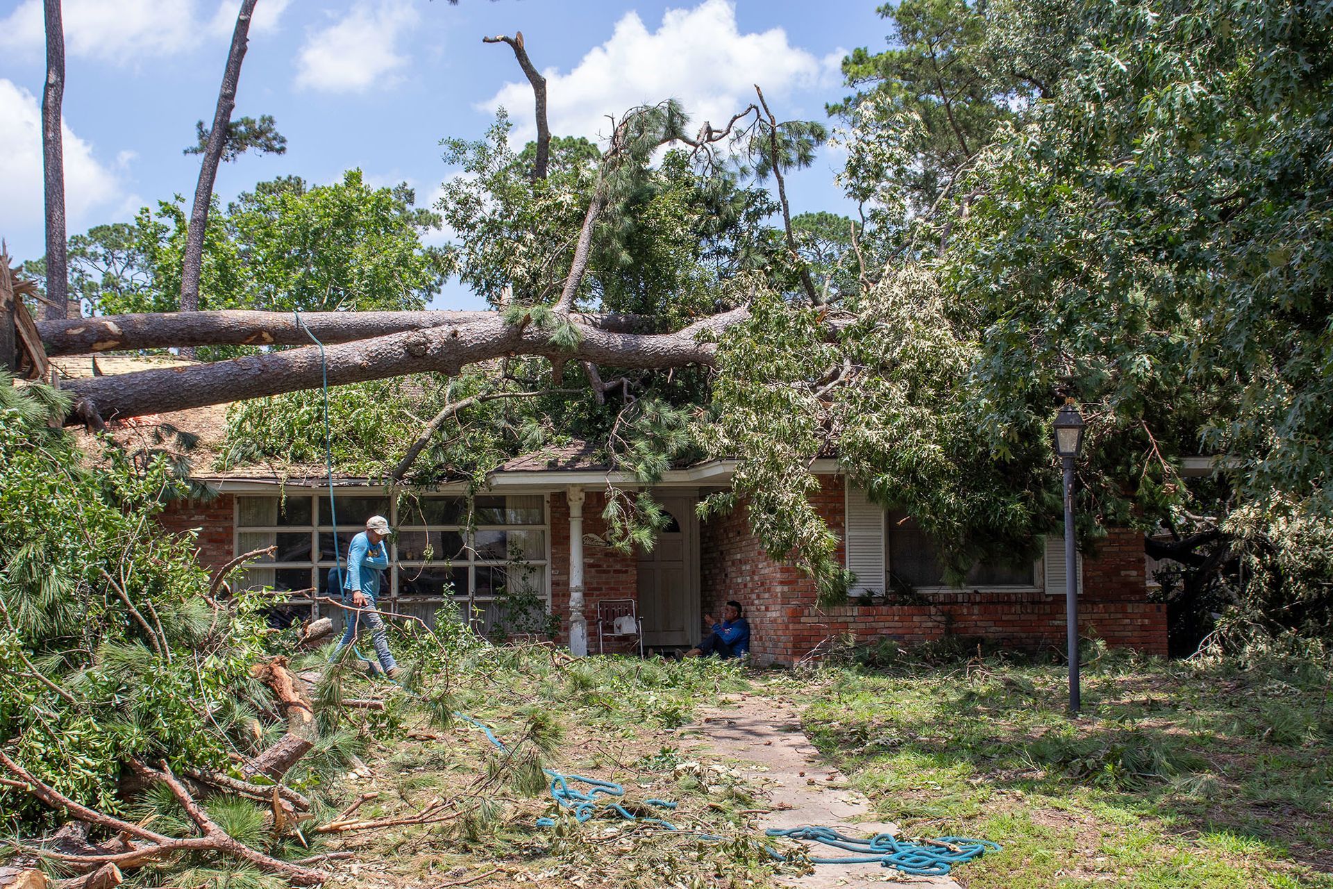 Storm-damaged yard with fallen tree across a brick house, debris scattered, and a person surveying the damage