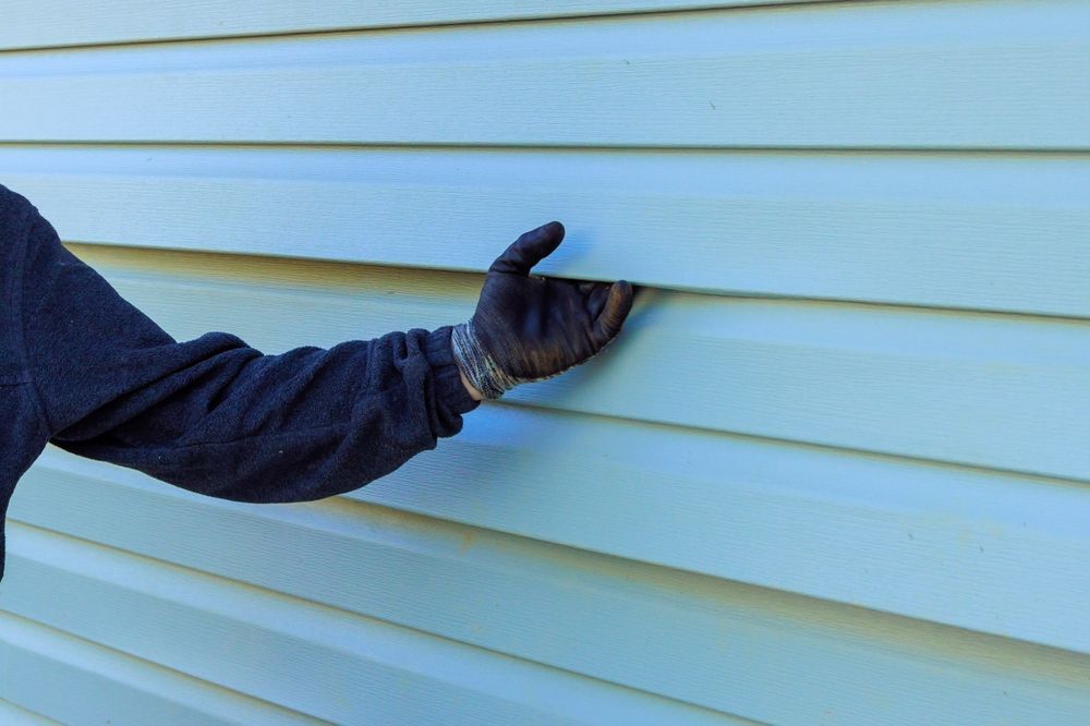Gloved hand prying at light blue vinyl siding on a building.