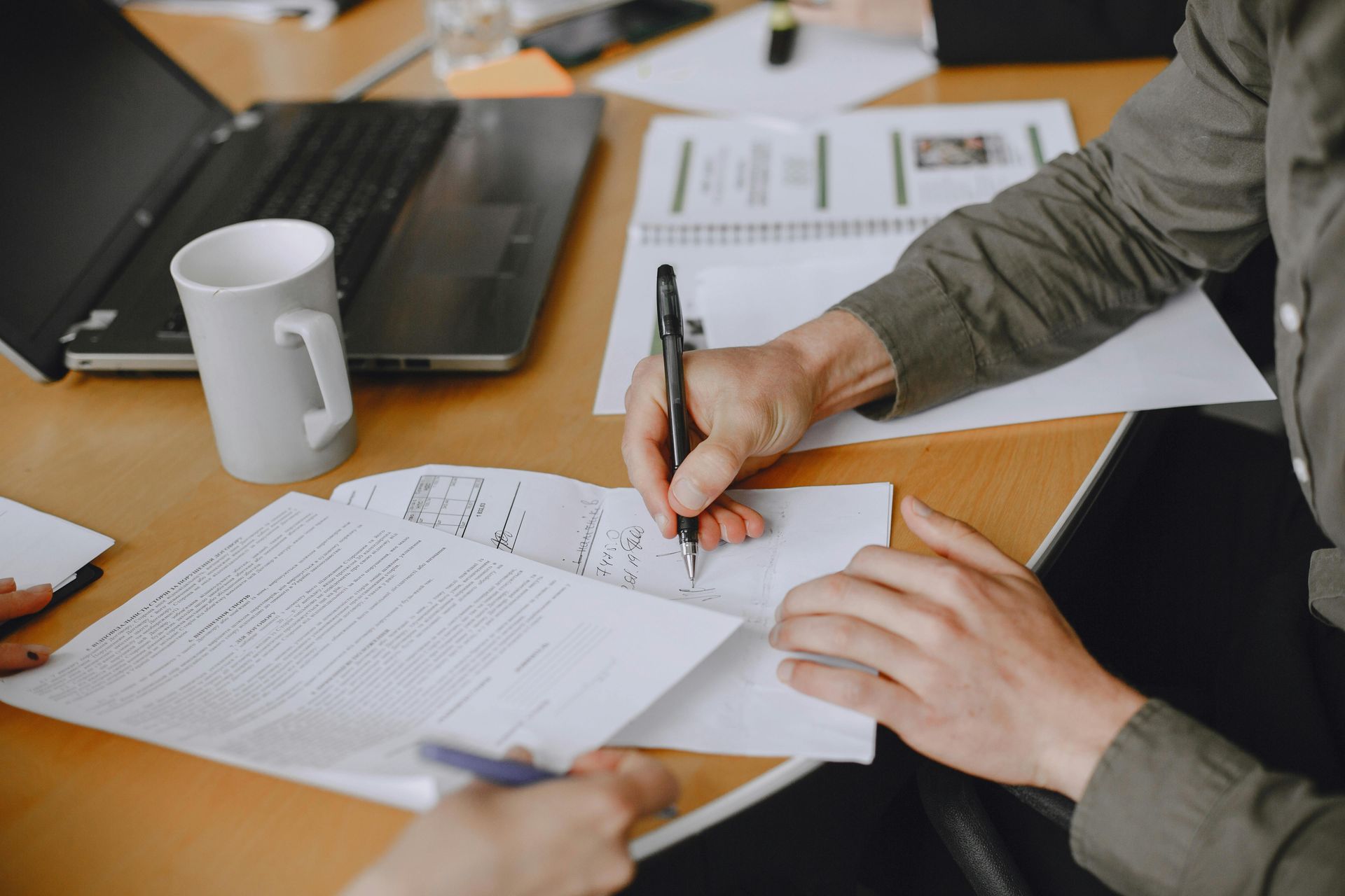 Hands writing on documents at a table, alongside a laptop, mug, and notebook.