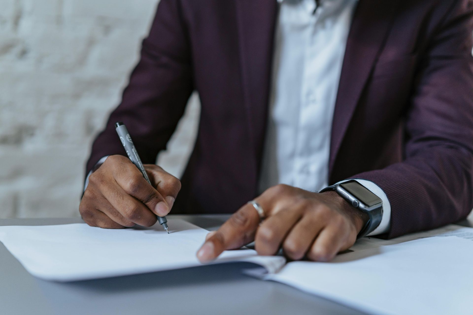Person in a suit writing on paper with a pen, pointing with the other hand.