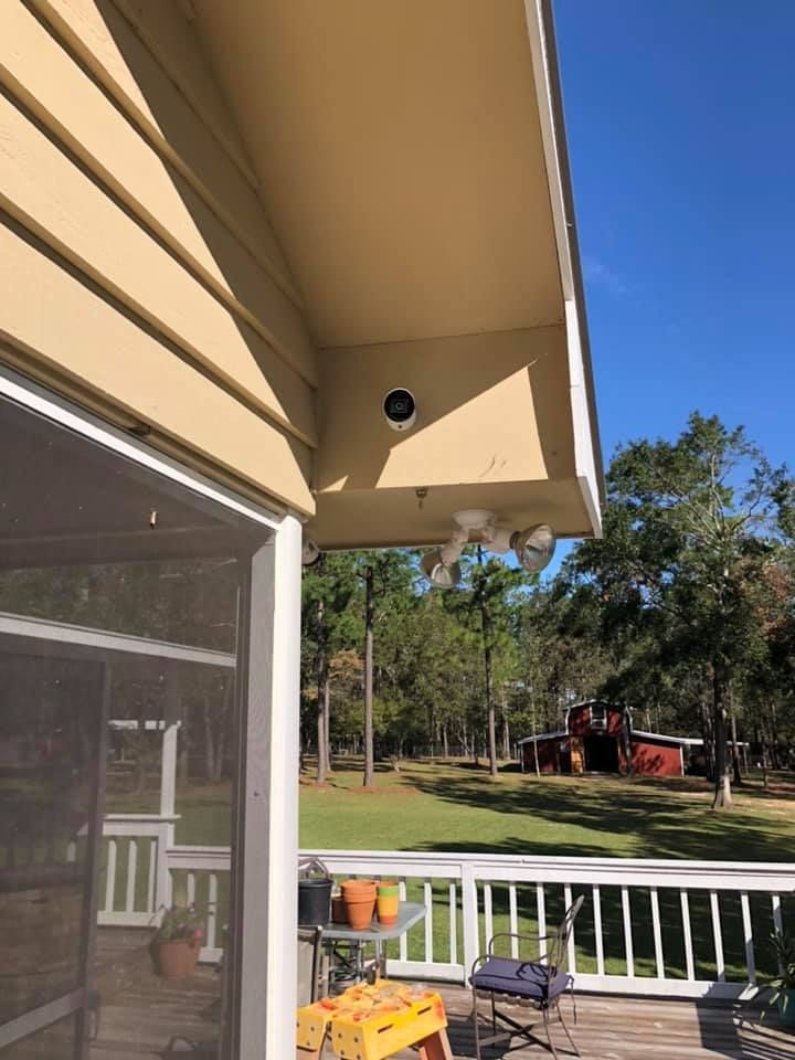 Overhang of a house with a security camera, porch with a white railing, trees, and a red building in the distance.