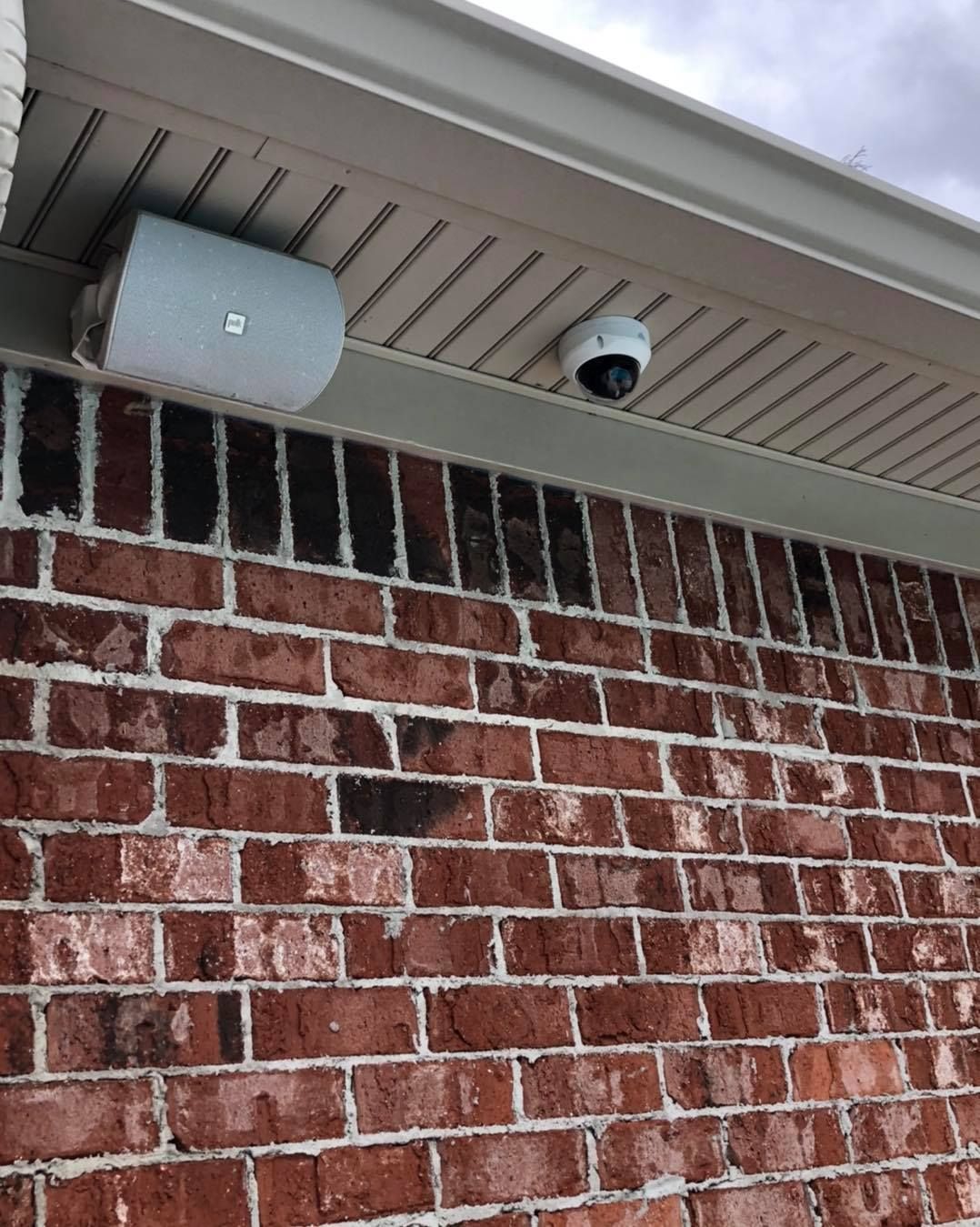Brick wall with a white security camera and speaker mounted under the eaves of a house.