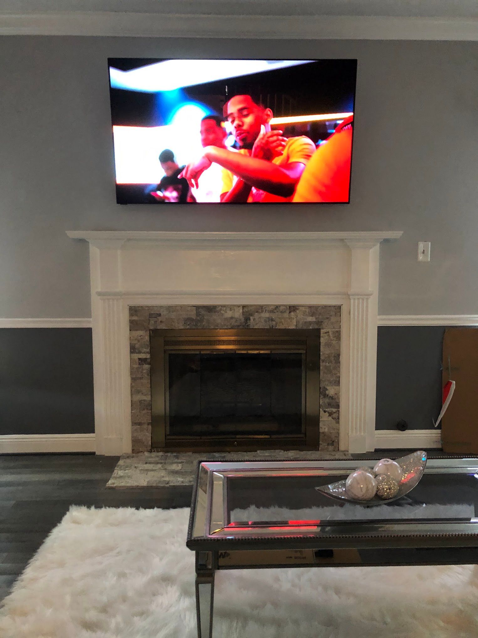 A TV above a fireplace, showing a person in an orange shirt. Gray walls and a dark-colored coffee table.