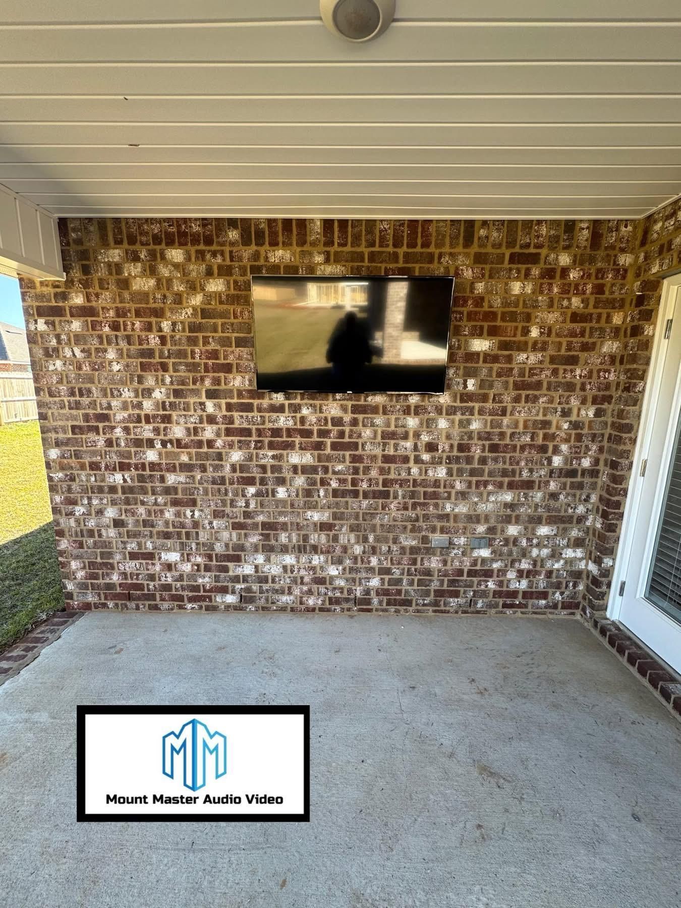 Outdoor TV mounted on a brick wall. A speaker is above the screen. Concrete patio.