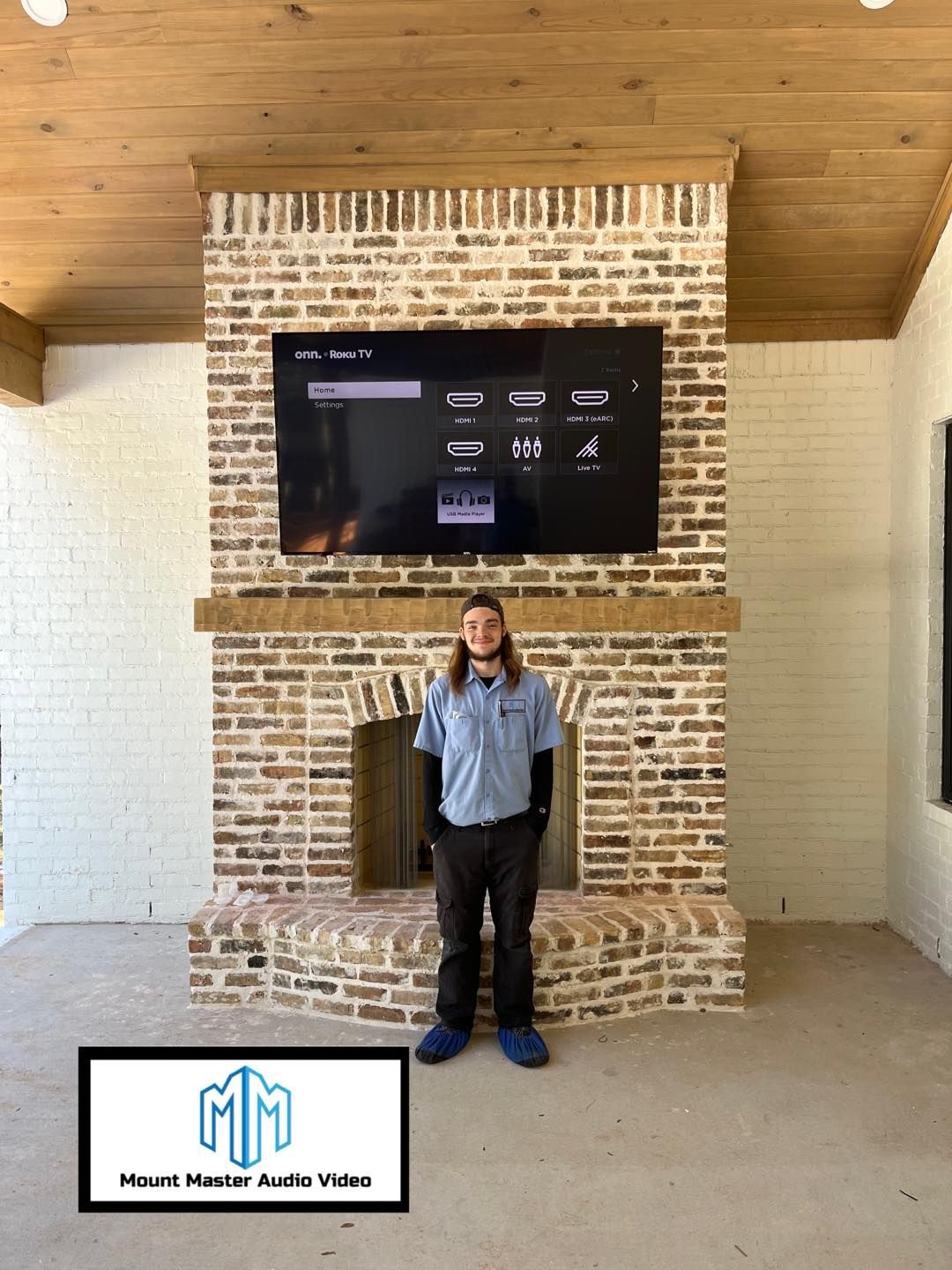 Man stands in front of a brick fireplace with a mounted TV. The room has wood beams and a white wall.