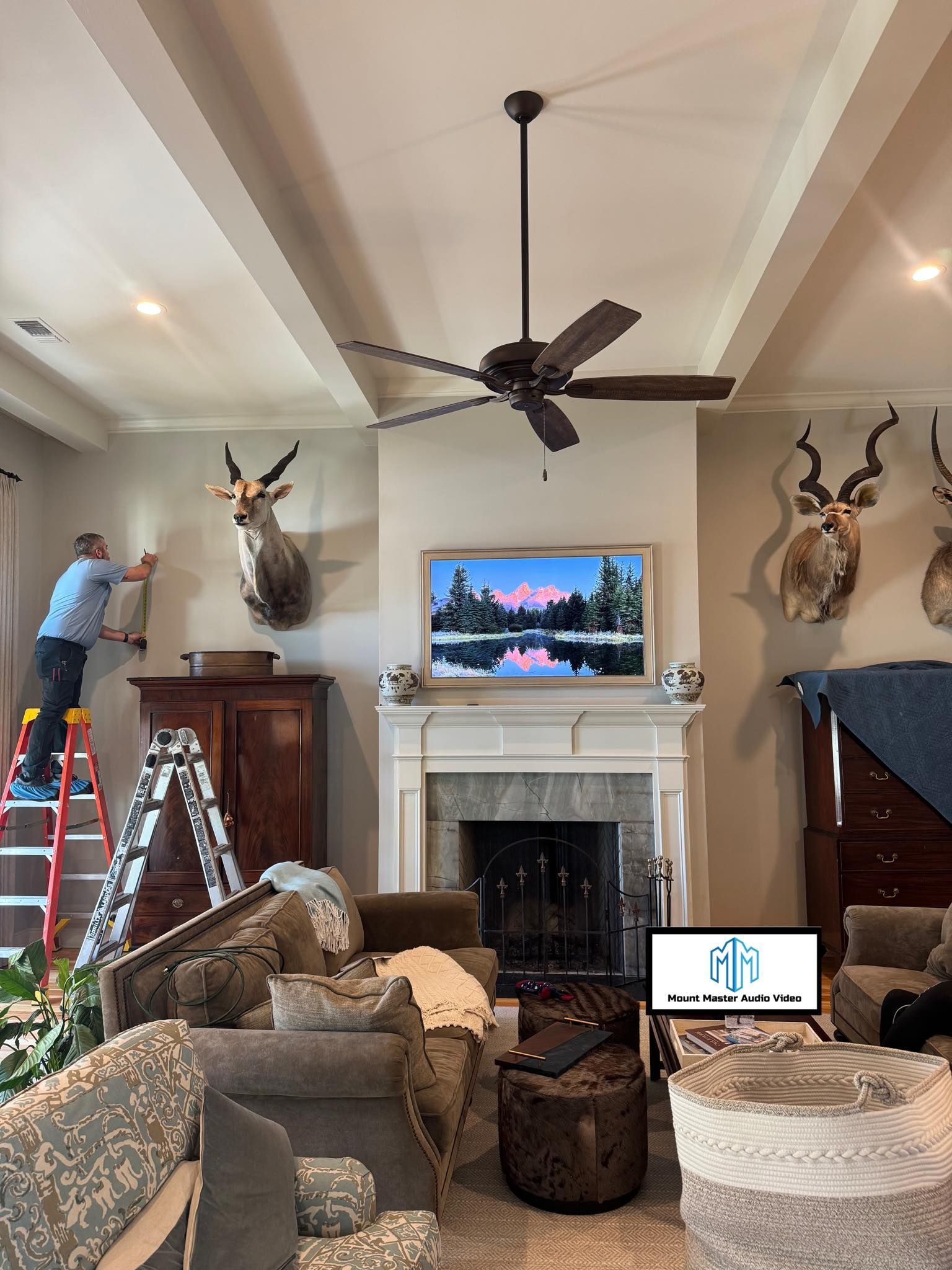Man on ladder adjusts a trophy on a wall in a living room with a TV and ceiling fan.