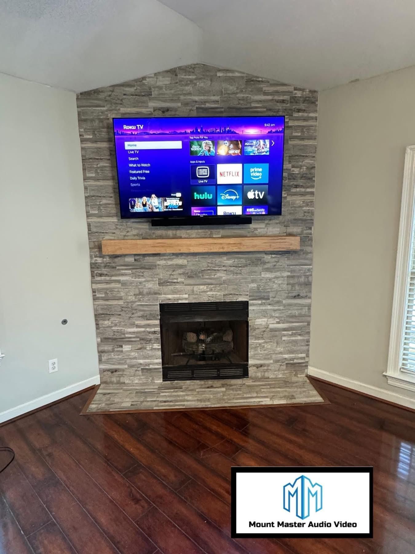TV above a fireplace, mounted on gray stone with a wooden beam mantle. Dark wood floor.