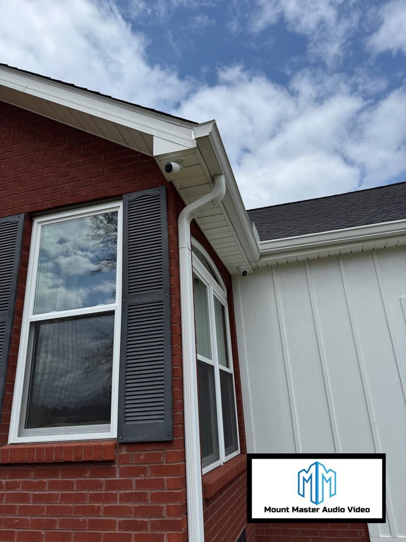 Red brick house with white trim, gutters, and a security camera. A window with gray shutters. Blue sky.