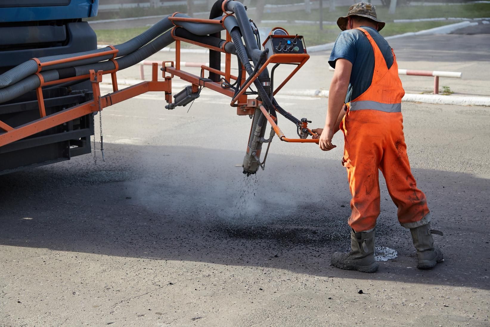 A road worker in orange overalls uses a truck-mounted sprayer to repair a pothole in the asphalt.