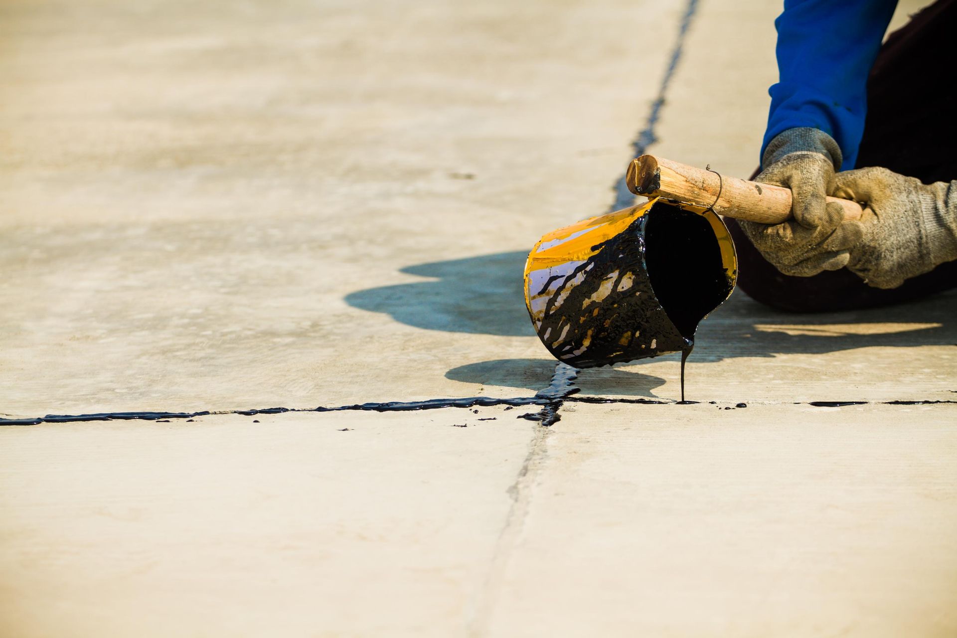 A person in work gloves pours black sealant from a yellow container into a crack in a concrete surface.