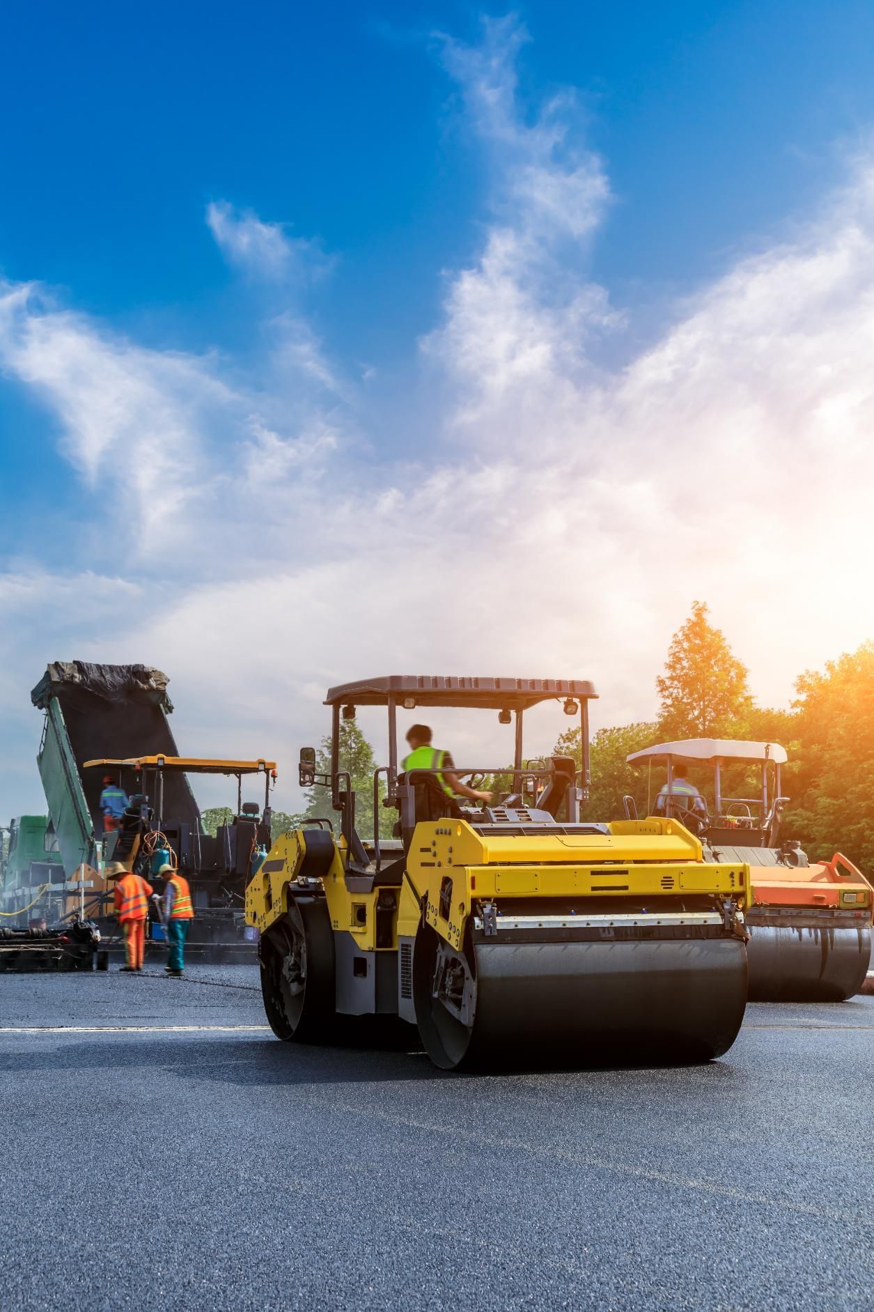 Construction workers operate yellow rollers and paving equipment on a road under a bright, sunny sky.