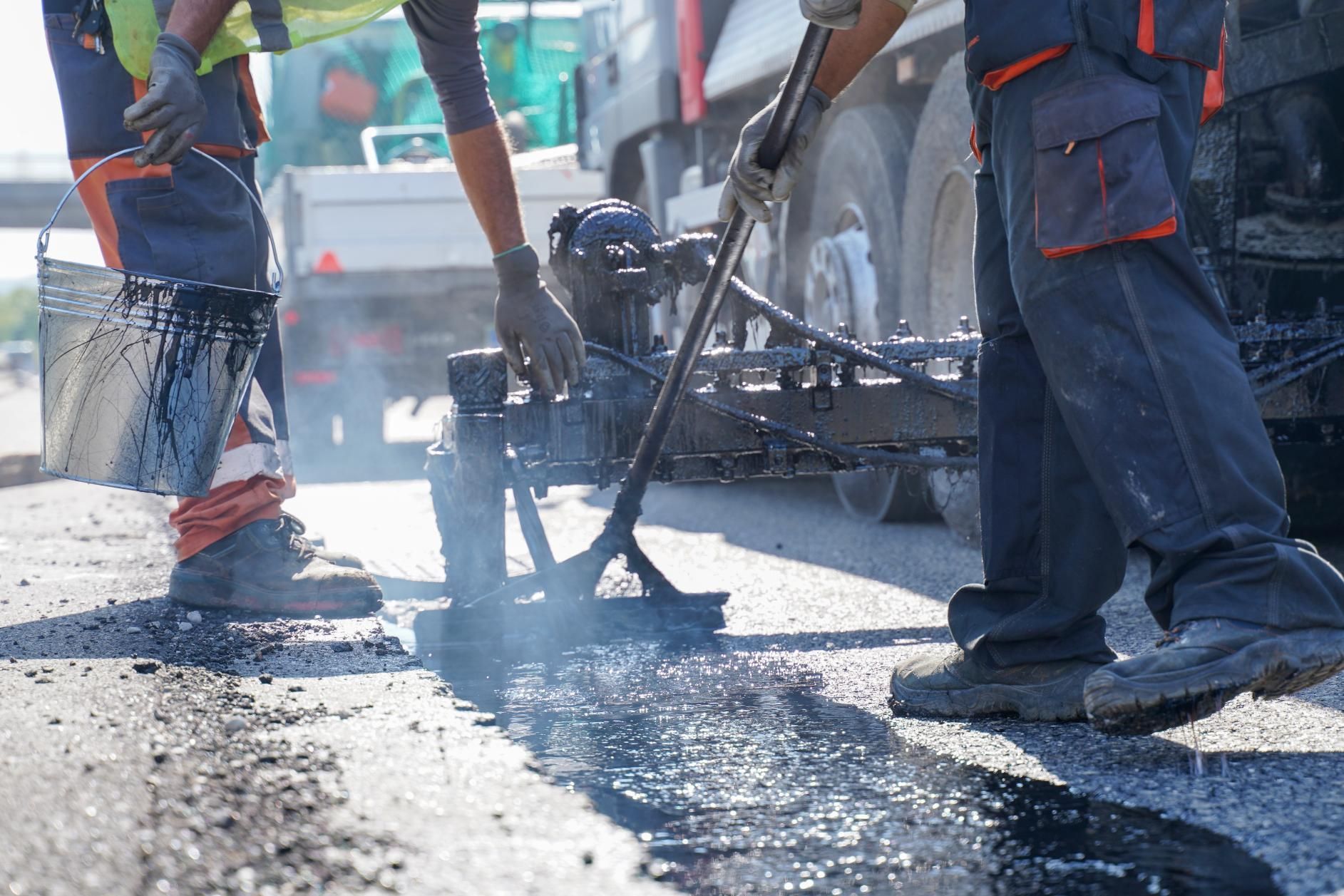 Two road workers in work gear use a squeegee and bucket to apply black sealant to an asphalt road surface.