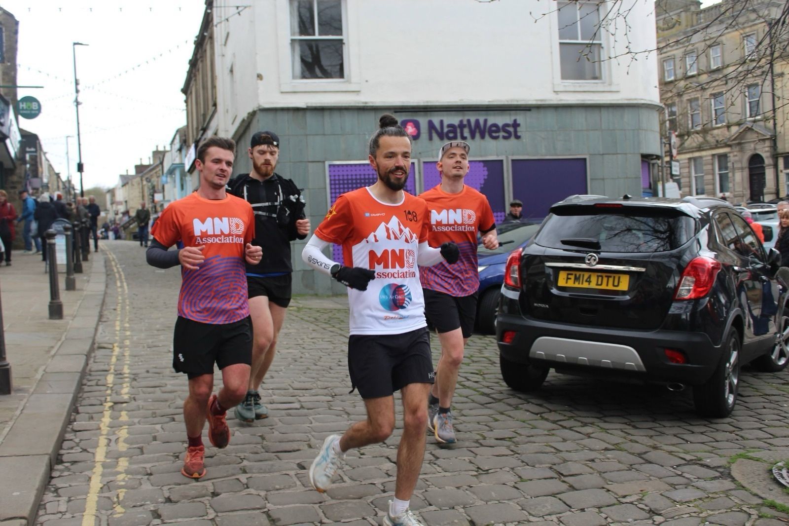 Runners in orange race shirts jog down a cobblestone street past a NatWest bank and parked cars.