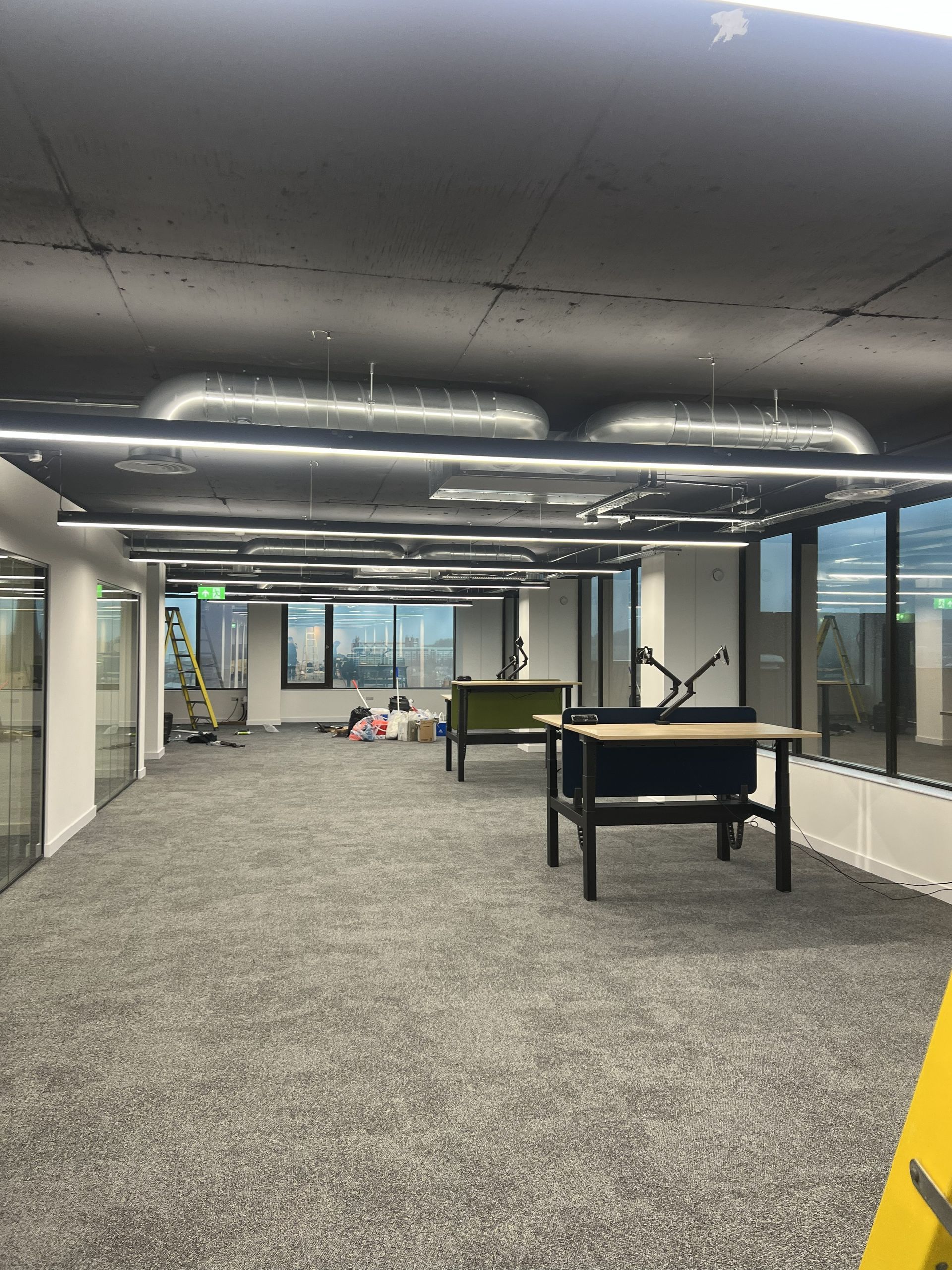 Modern office lounge with carpet, desks, overhead lights, and large windows overlooking a city view