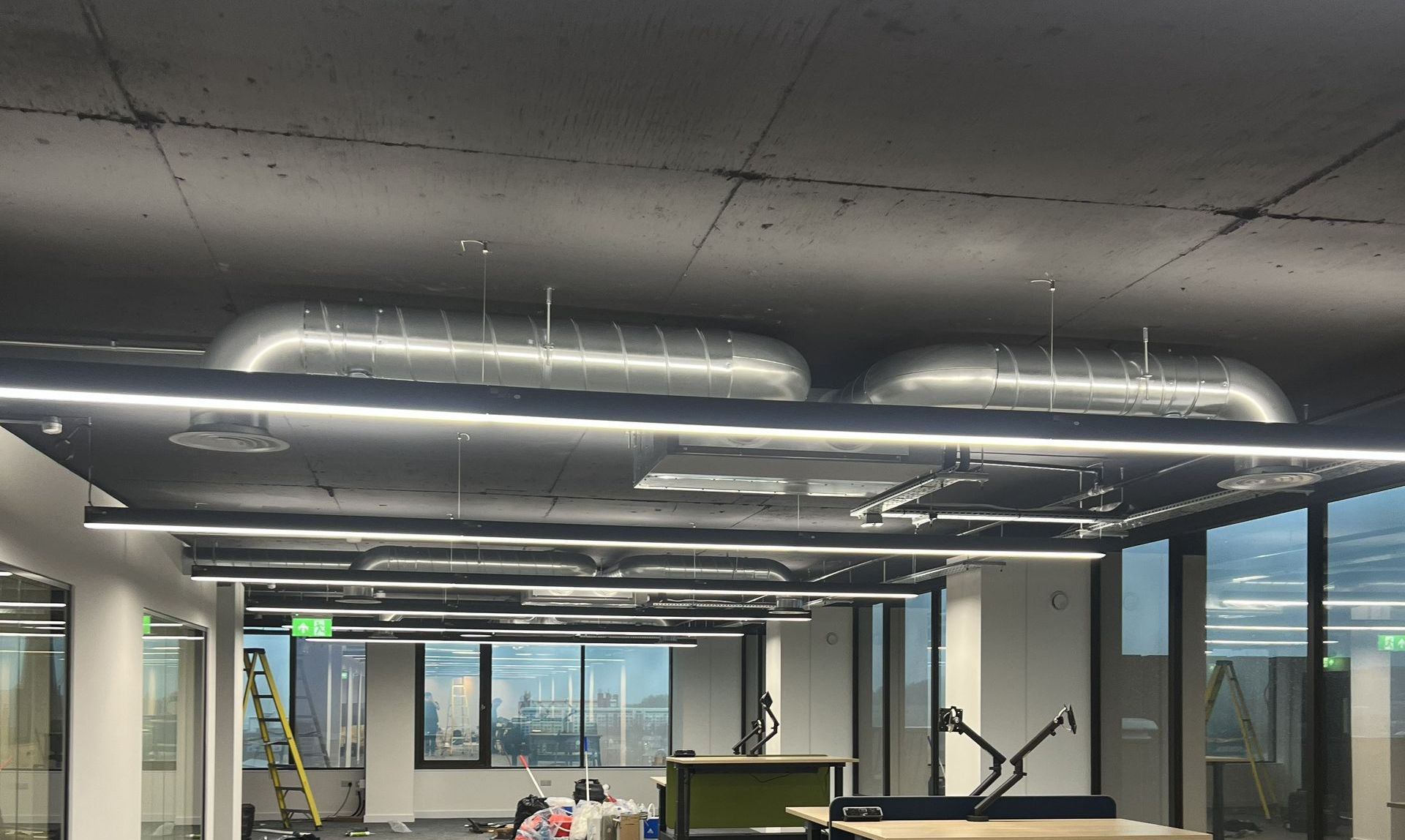 Modern office interior with exposed ceiling lights, desks, and large windows overlooking trees