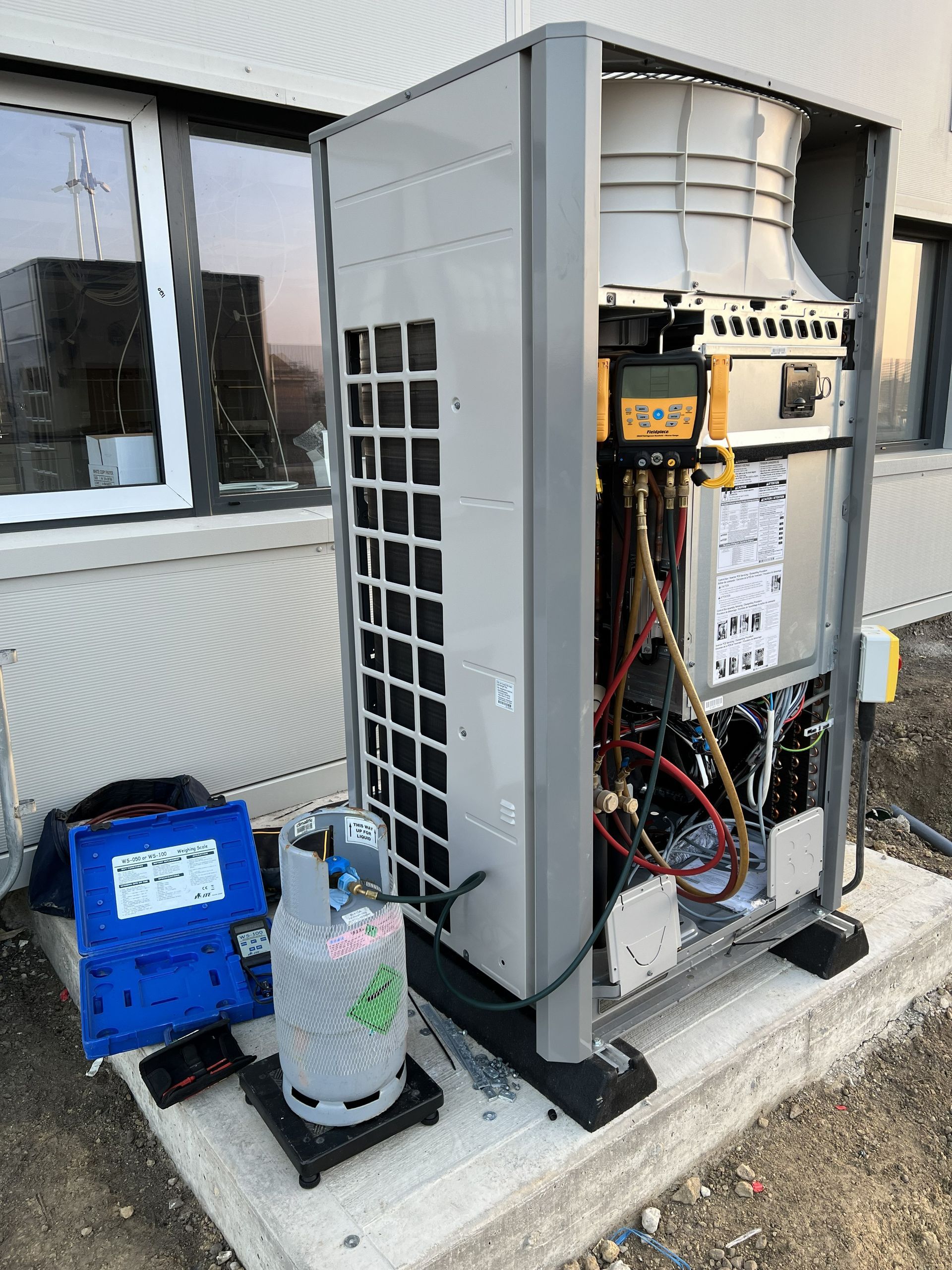Open electrical service cabinet beside a building, with exposed wiring and blue equipment on the ground.