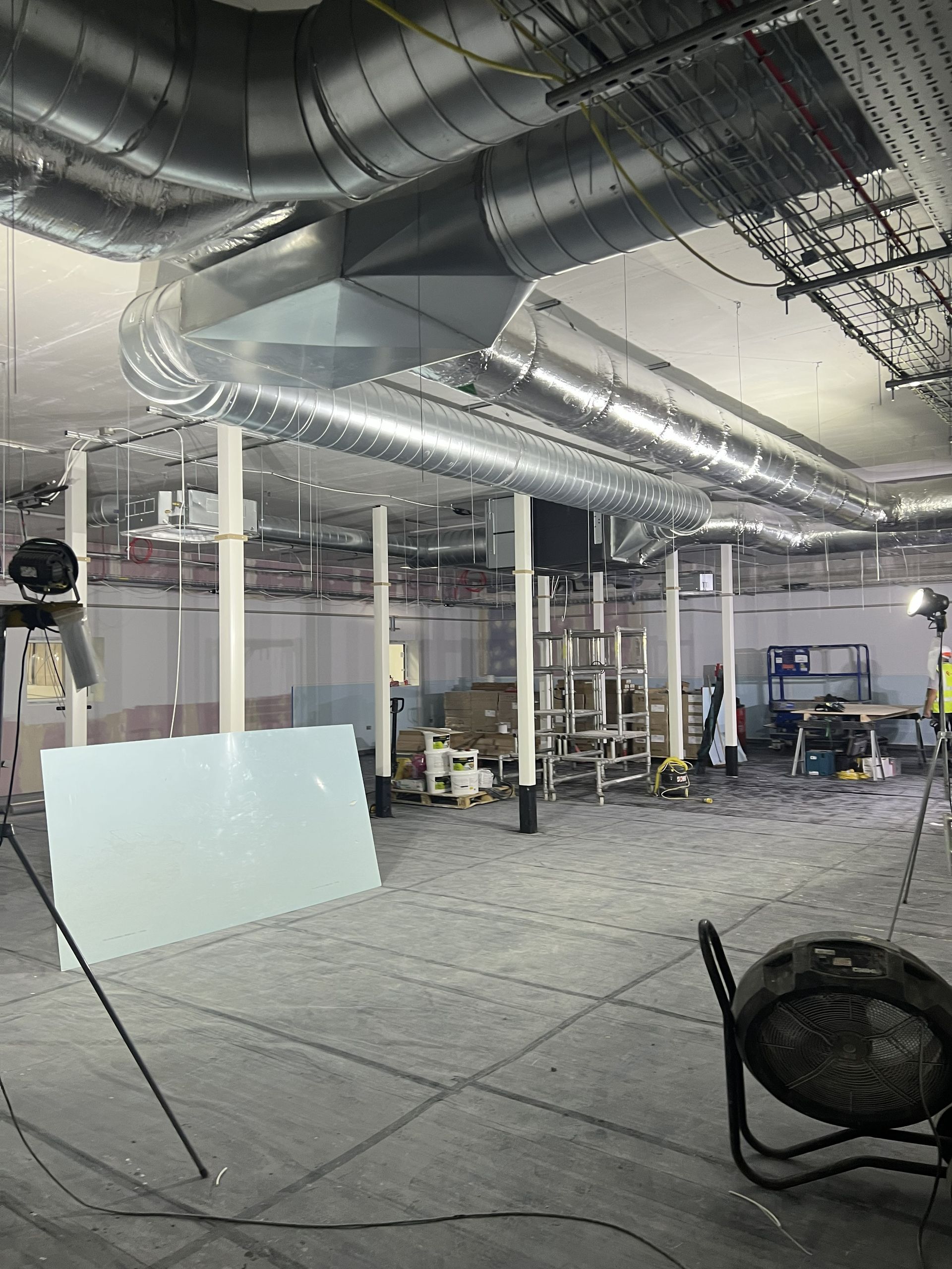 Partially finished industrial interior with exposed ducts, white columns, scattered tools, and construction materials
