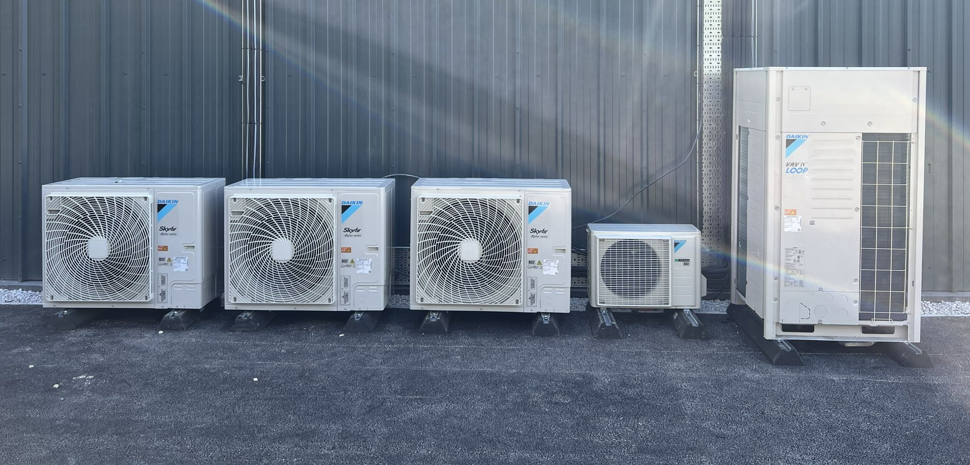 Four outdoor HVAC units lined up on gravel against a corrugated metal wall