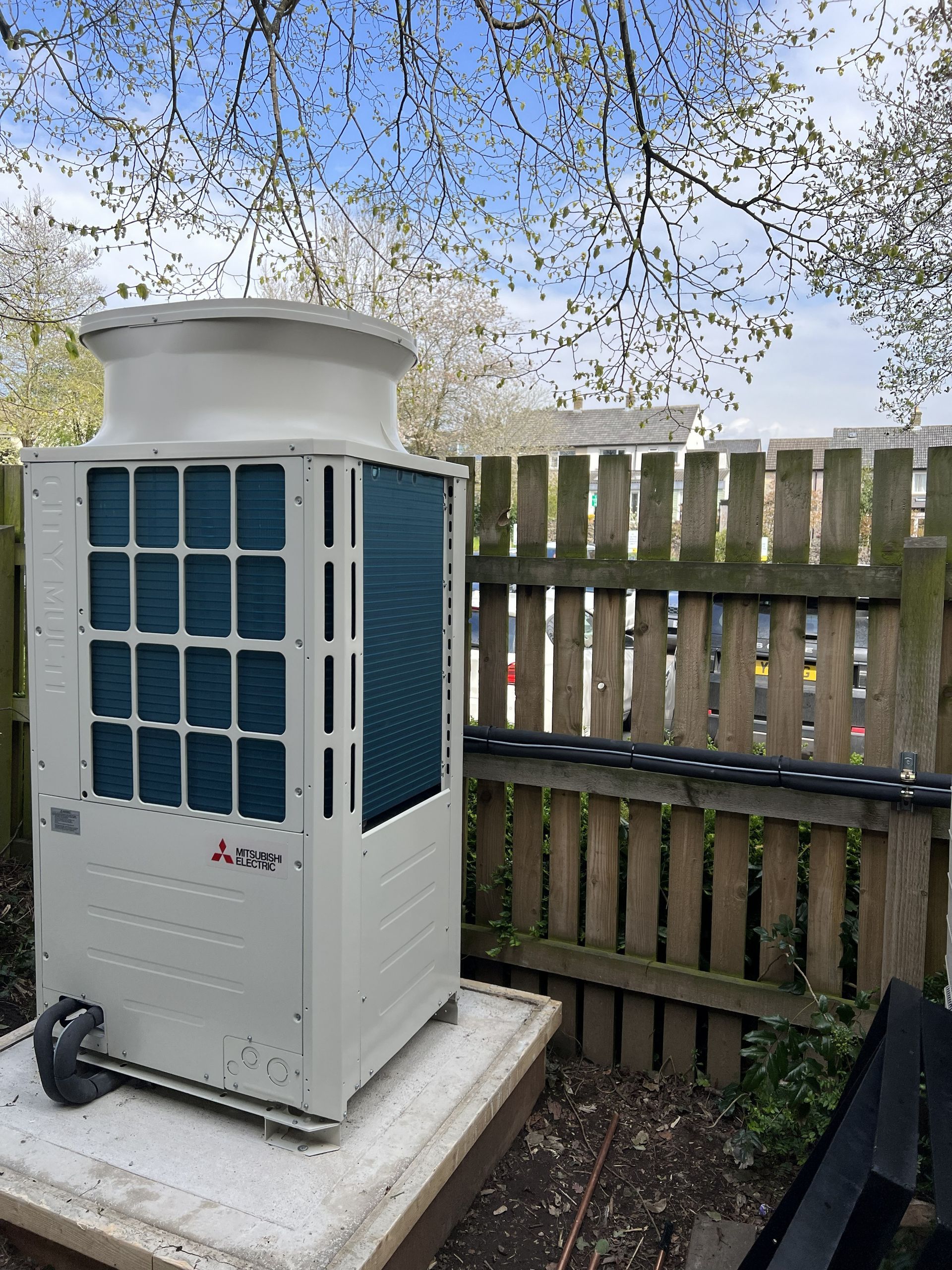 Outdoor air conditioner unit beside a wooden fence under a tree canopy