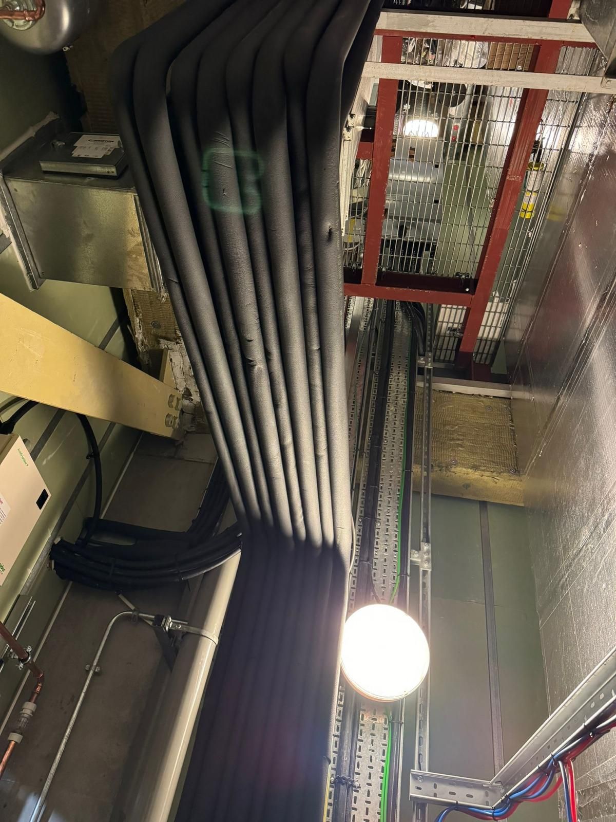 Ceiling with bundled black cables, red mesh guard, and a bright hanging light in an industrial space