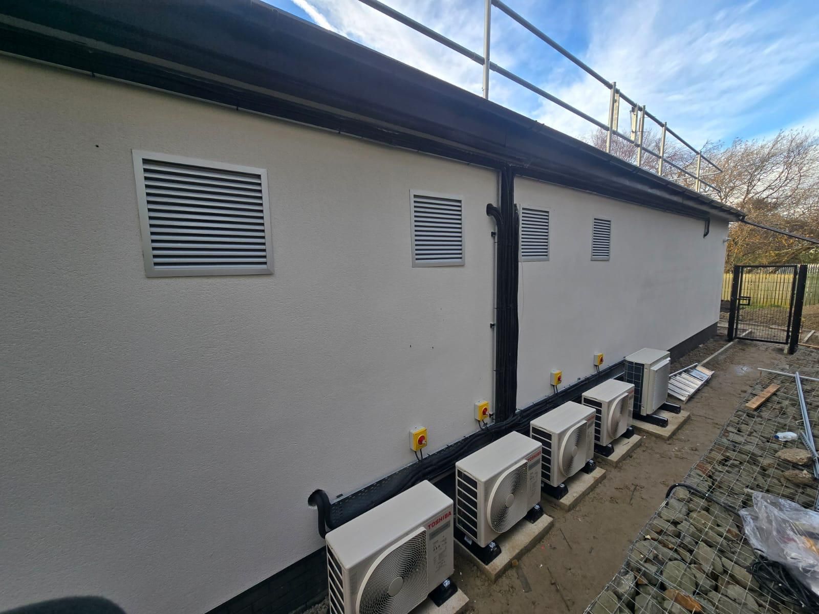 Row of outdoor HVAC units beside a beige building with vents and a fenced side yard