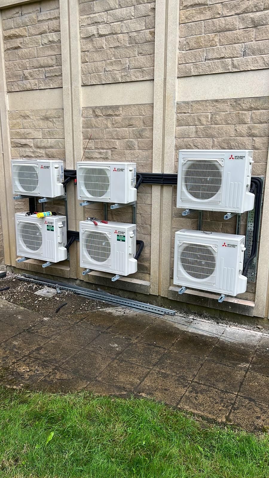 Six outdoor air conditioner units mounted on a stone wall beside a grassy strip