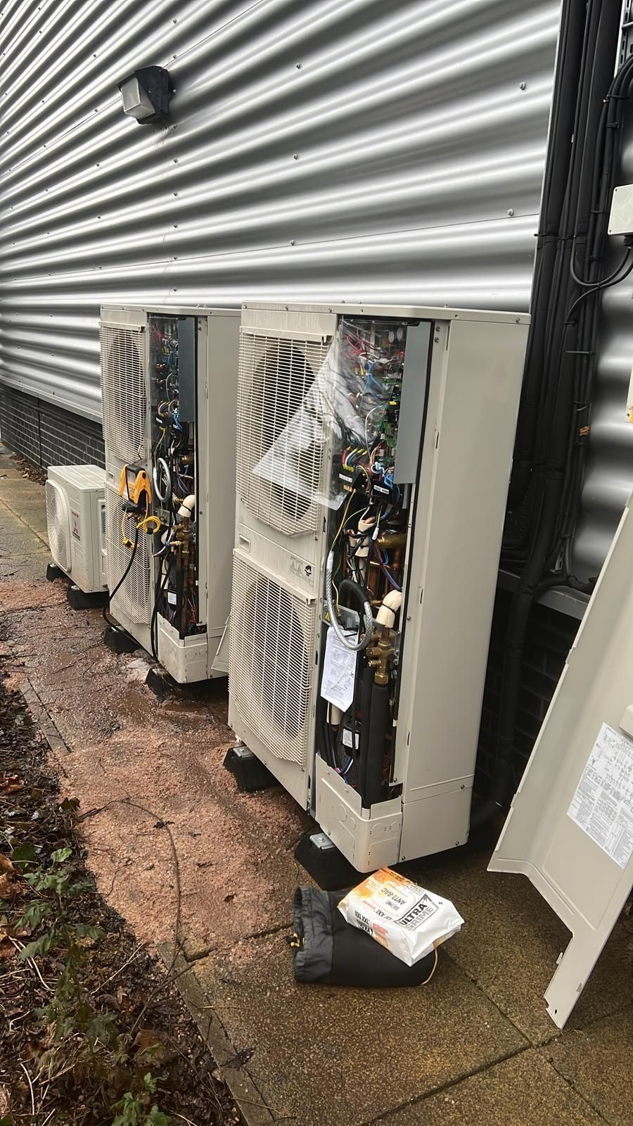 Outdoor HVAC units beside a corrugated metal building, with exposed wiring and open panels on a wet walkway.