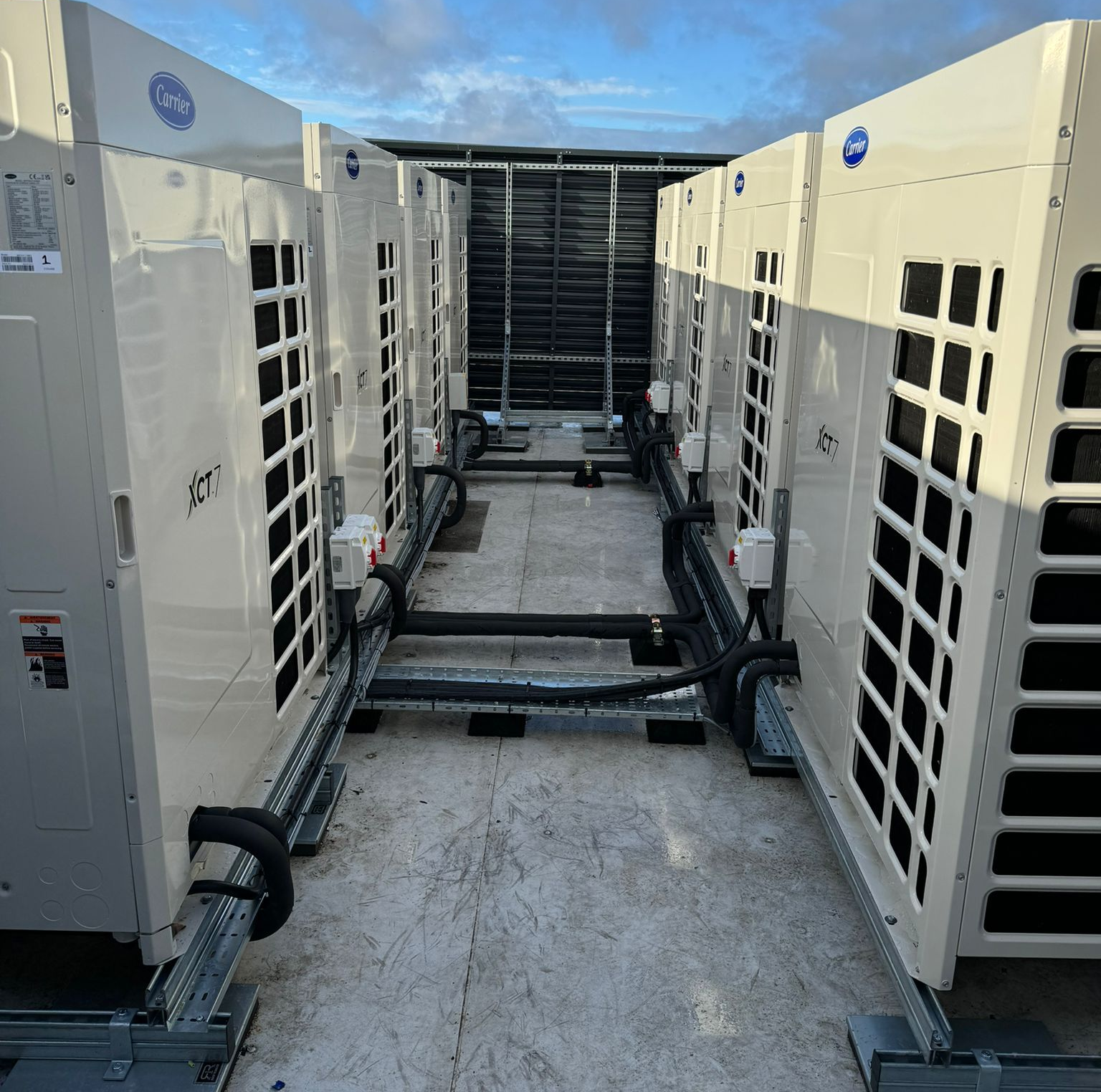 Outdoor row of large white HVAC units on a rooftop, with pipes and cables between them under a blue sky
