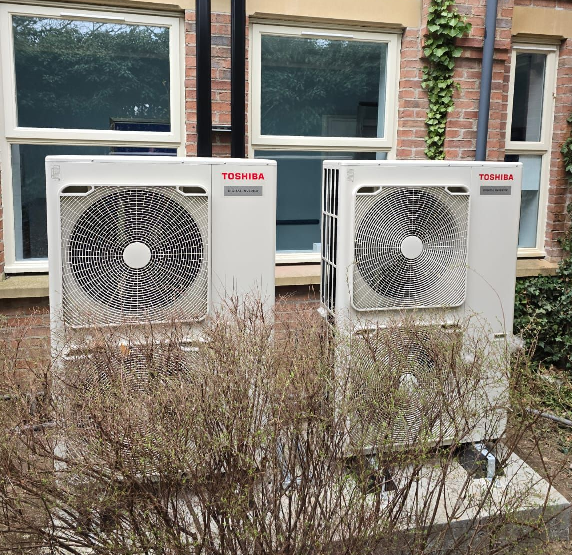Two white outdoor AC units beside a brick building, with windows and overgrown shrubs in front.