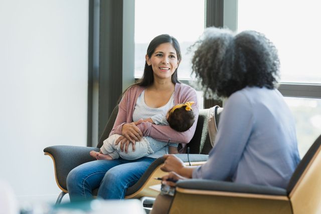 A woman is sitting in a chair holding a baby and talking to another woman.