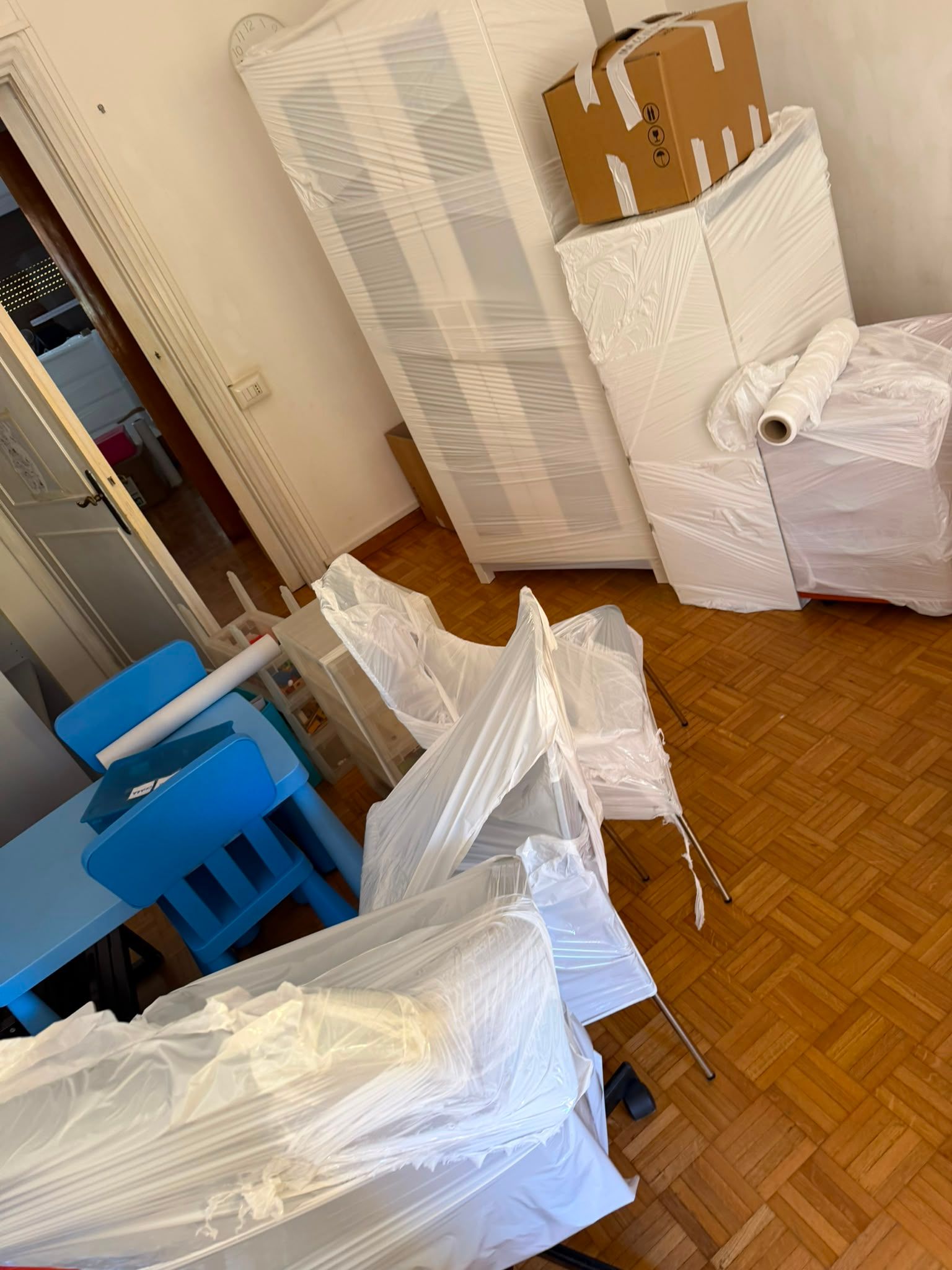 Stanza stipata con mobili rivestiti, sedie blu e scatole di cartone impilate su un pavimento di legno.