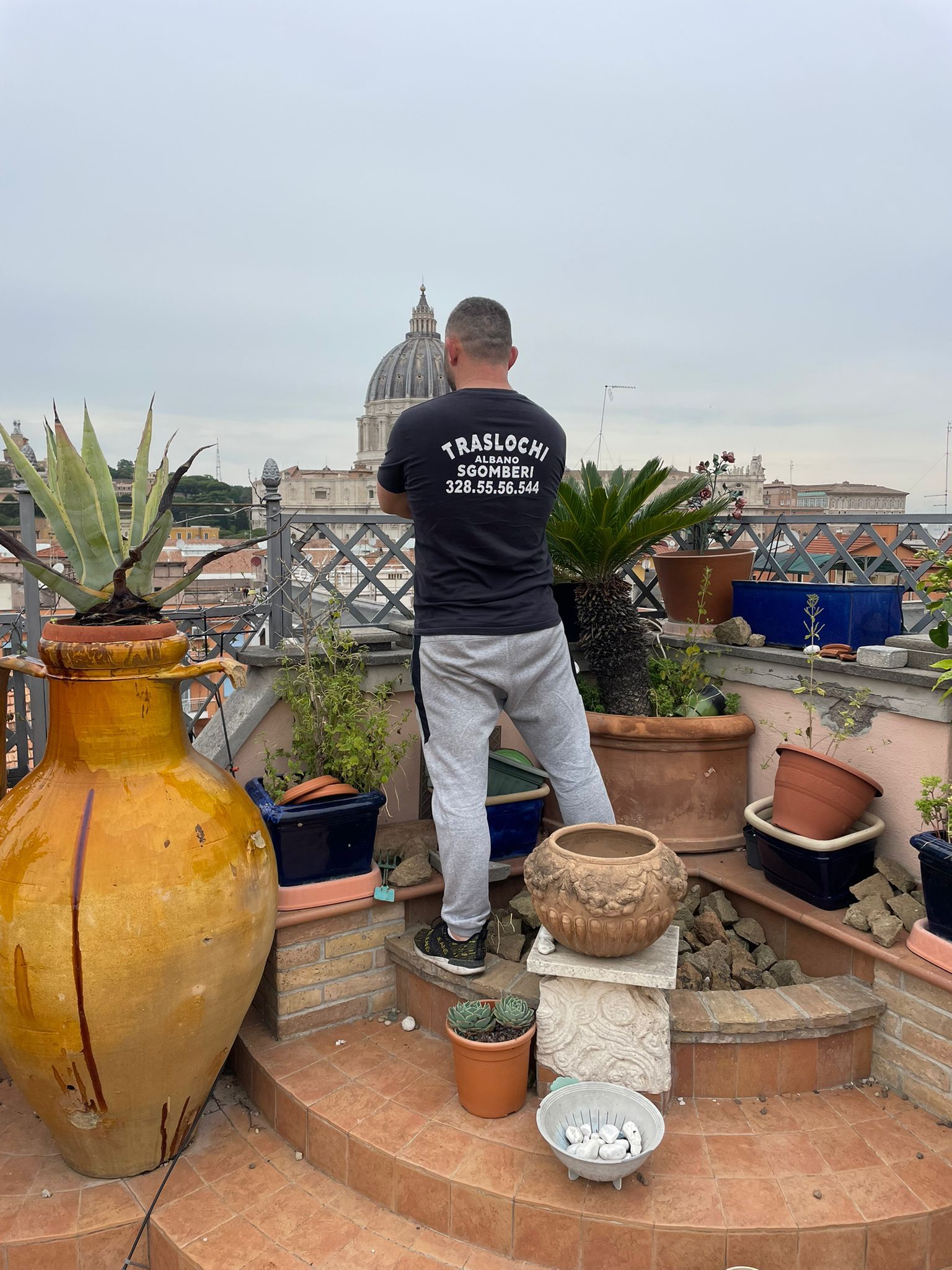 Un uomo in piedi su una terrazza sul tetto, con piante in vaso, guarda lo skyline della città sotto un cielo nuvoloso.