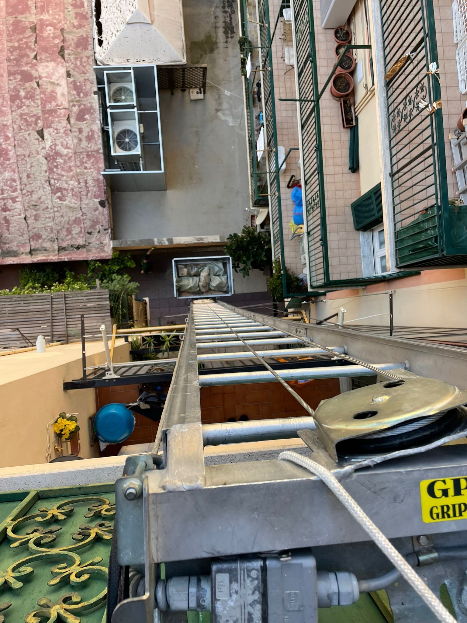 Uno sguardo giù da una stretta scala tra gli edifici verso un tetto con tubi, cavi e apparecchiature di ventilazione.