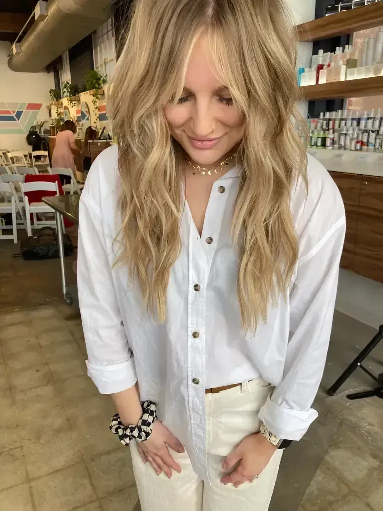 Person in a white shirt and jeans standing indoors, looking down with long wavy blonde hair.