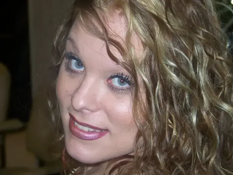Close-up of a smiling woman with wavy blonde hair and blue eyes, wearing pink lipstick.