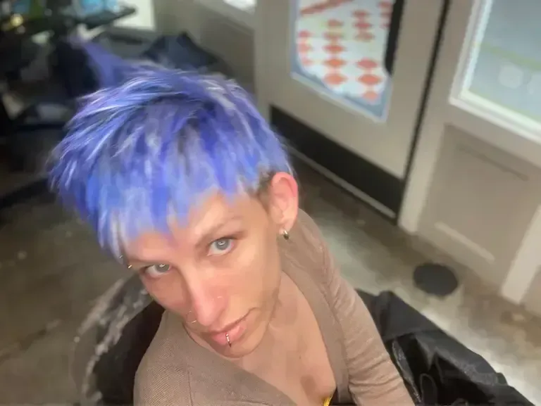 Person with bright blue hair looking up indoors, wearing a tan top and black jacket.