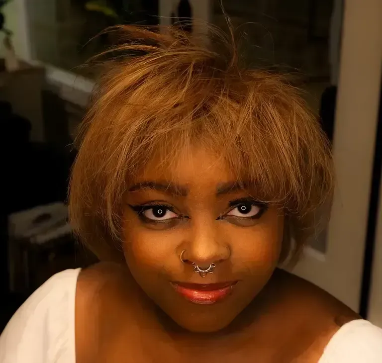 Portrait of a person with short blonde hair, nose ring, and white top, looking at the camera