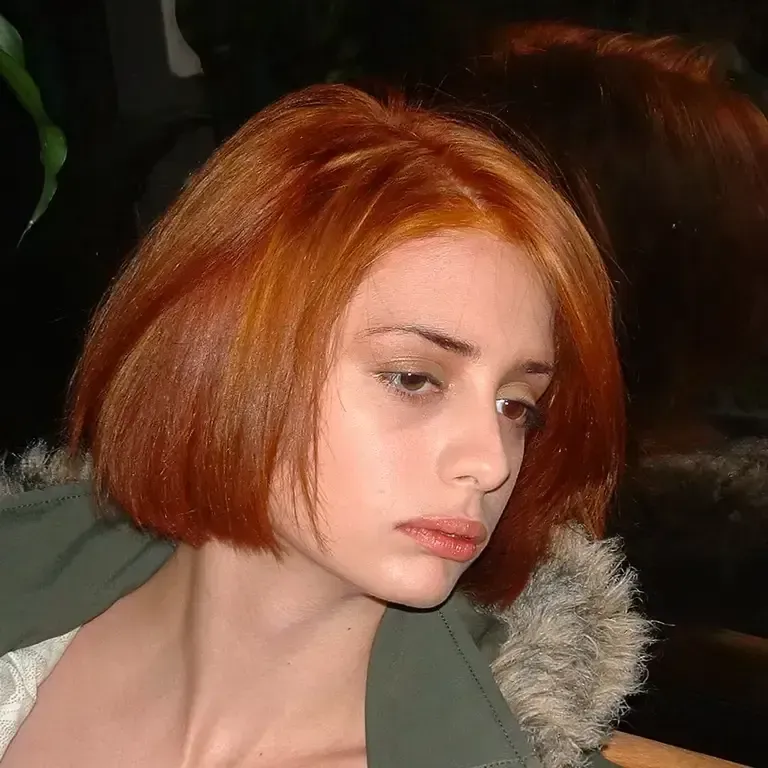 Portrait of a person with a short red bob haircut, looking down against a dark background