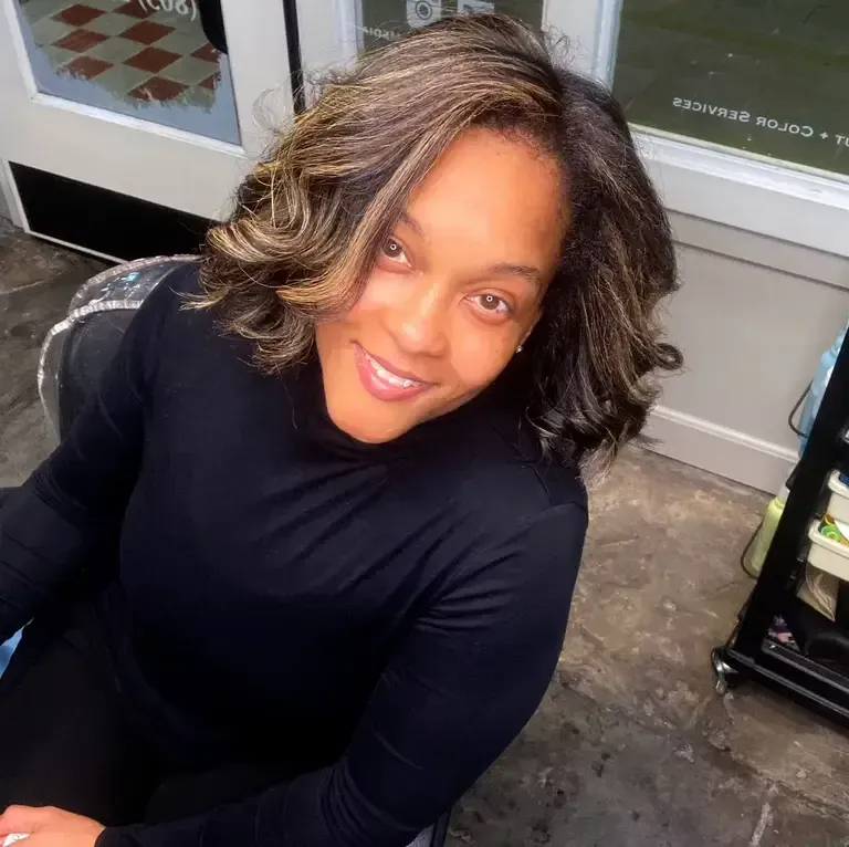 Smiling woman in a black top seated indoors near a glass door, looking up at the camera