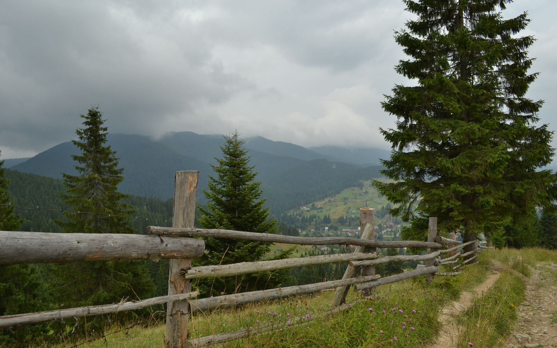 Wooden fence beside a grassy trail overlooking misty green mountains under cloudy skies
