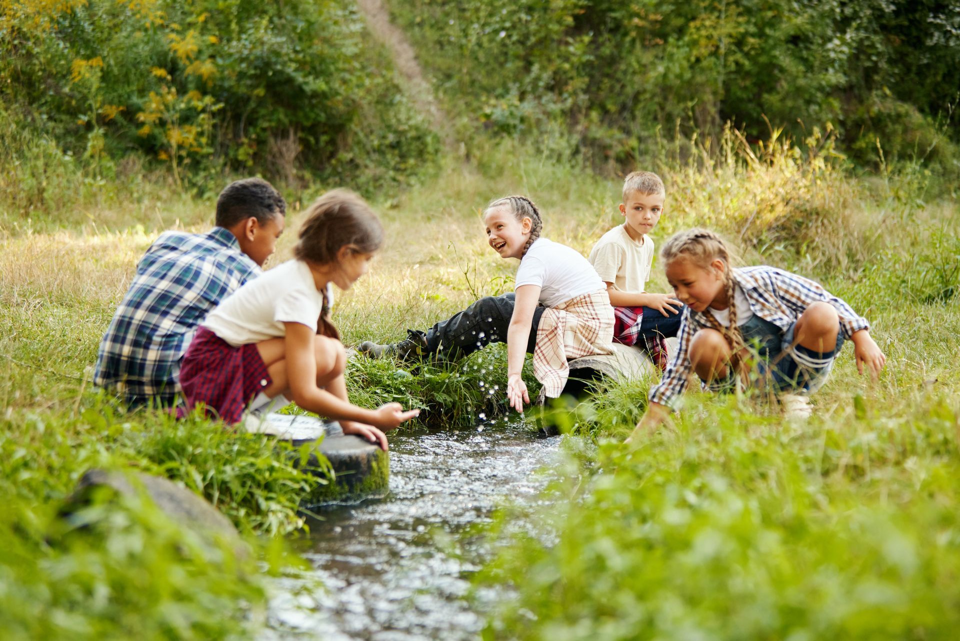Children crouch by a stream in a grassy field, washing hands and exploring nature together.